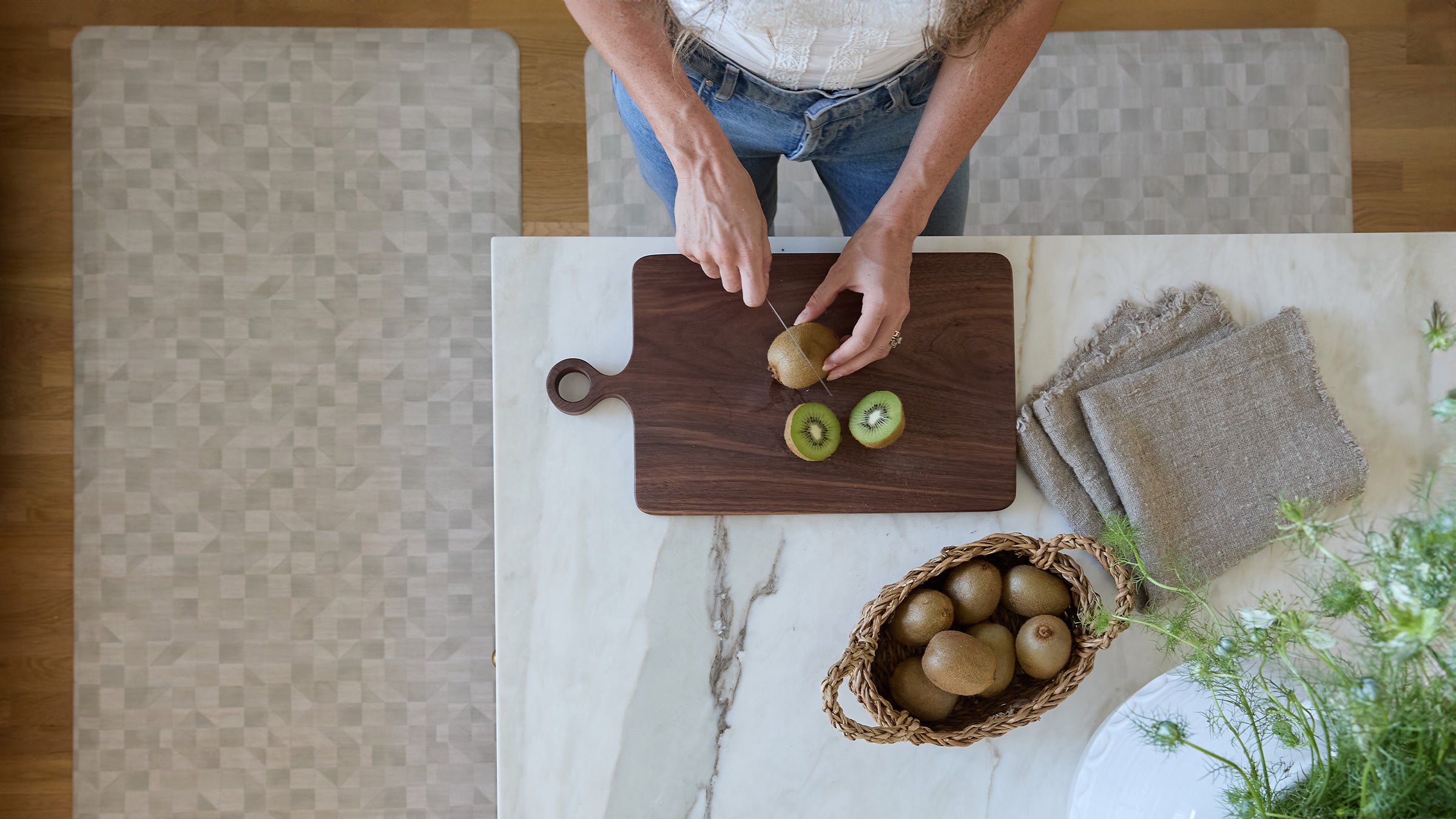 Aerial shot of the georgia clay neutral checker print standing mat in size 22x54 and 30x72 around a kitchen counter with a woman standing on size 22x54 cutting up kiwis 