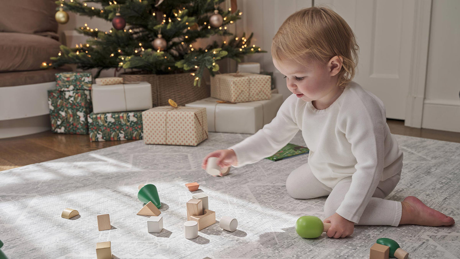 Ula gray play mat with toddler sitting on the mat playing with green and brown wooden blocks. Lit up christmas tree with gifts underneath in the background