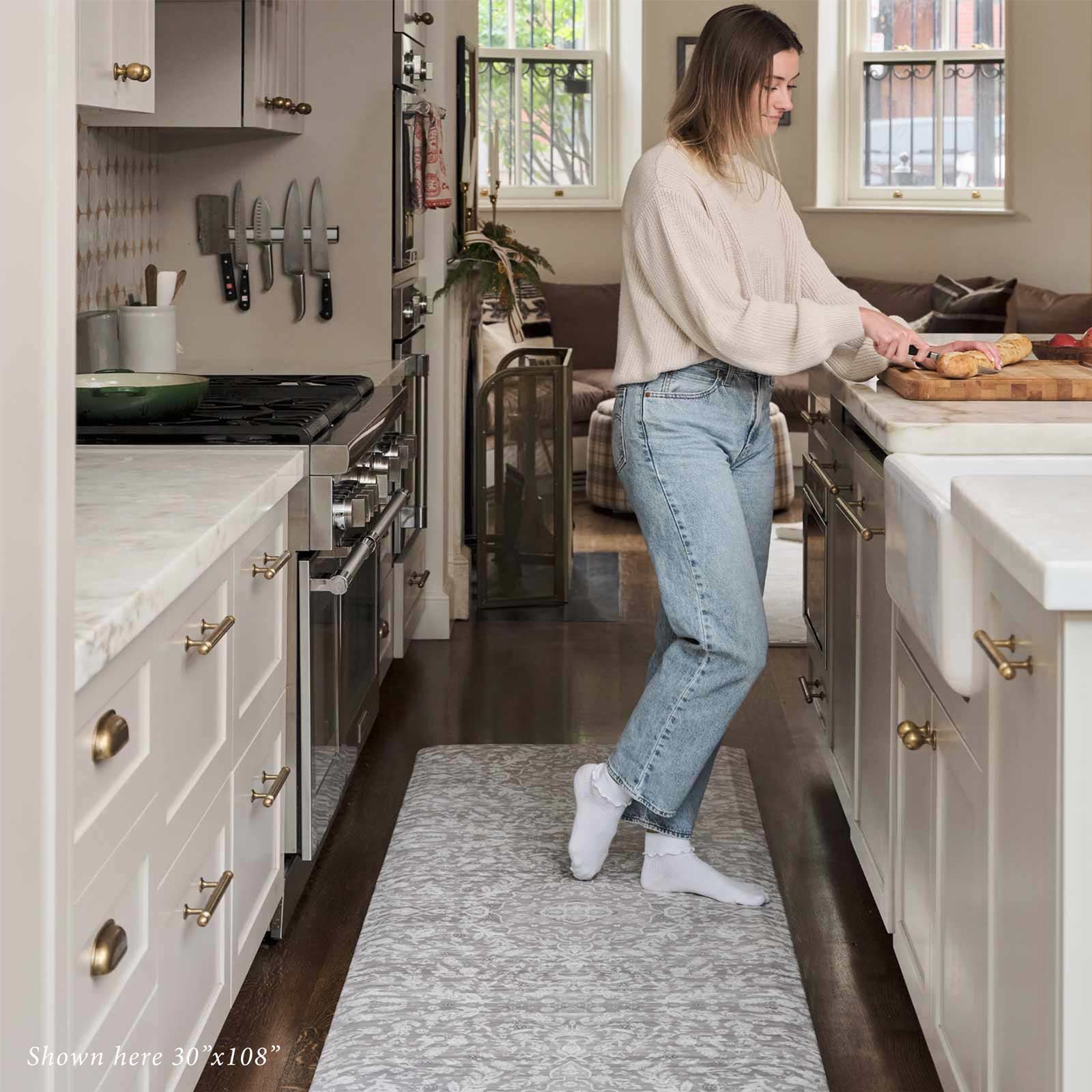 Emile earl grey gray and white floral standing mat shown in kitchen in size 30x108 with woman standing on the mat cutting bread on a cutting board on the counter