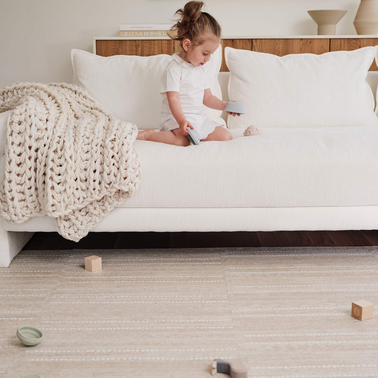 Aspen toast neutral minimal stripe little nomad play mat shown in a living room under a white couch with toddler sitting on the couch playing with blocks