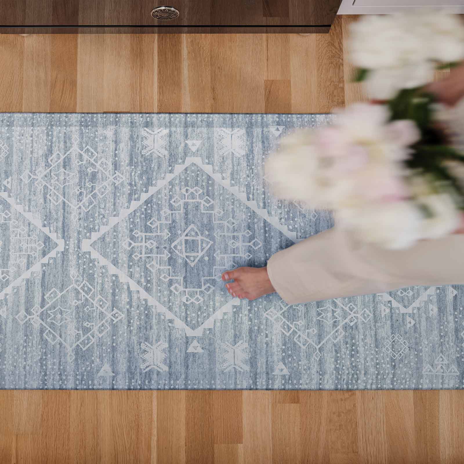 Ula blue and white boho print standing mat in size 30x72 shown from above on a wooden floor with a woman walking across the mat carrying white flowers