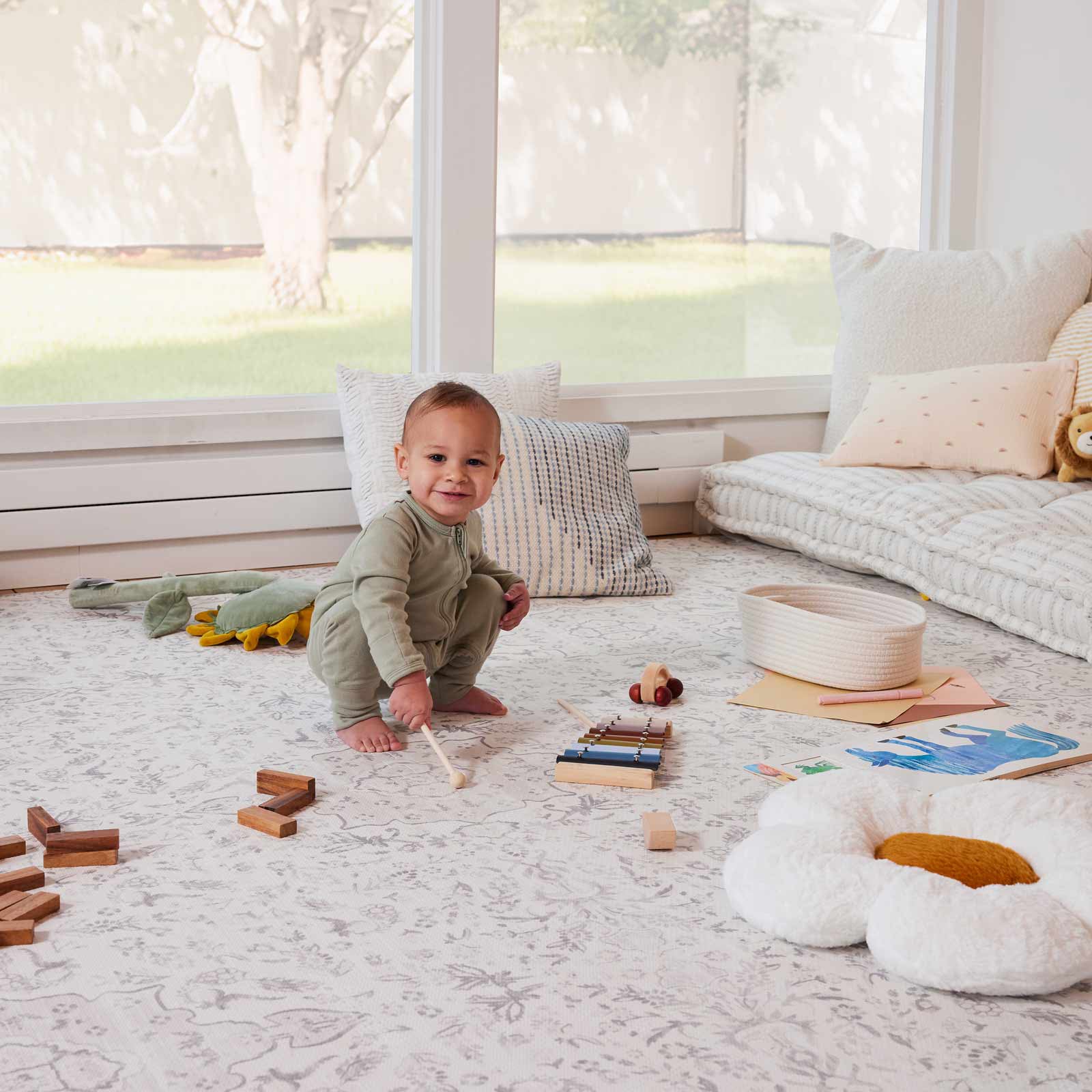 Emile Latte neutral floral little nomad play mat shown in a play room with baby boy smiling surrounded by toys and plush pillows on the mat