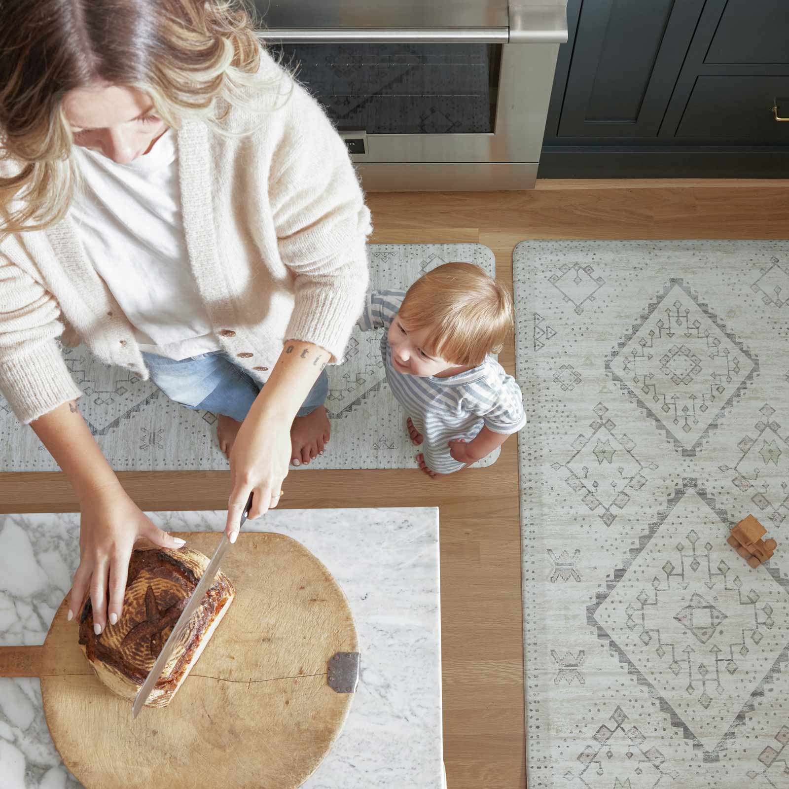 Ula Calico neutral boho print standing mat shown from above in sizes 22x72 and 30x72 surronding a kitchen counter with a woman standing at the counter cutting bread