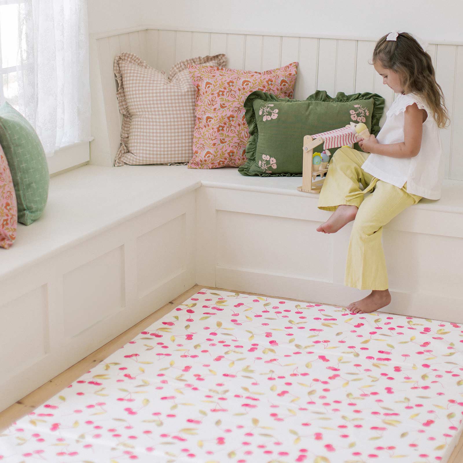 Lulu red and green Cherry print tumbling mat shown on the floor in front of a corner window seat filled with pink and green pillows and little girl sitting on the seat playing with a toy