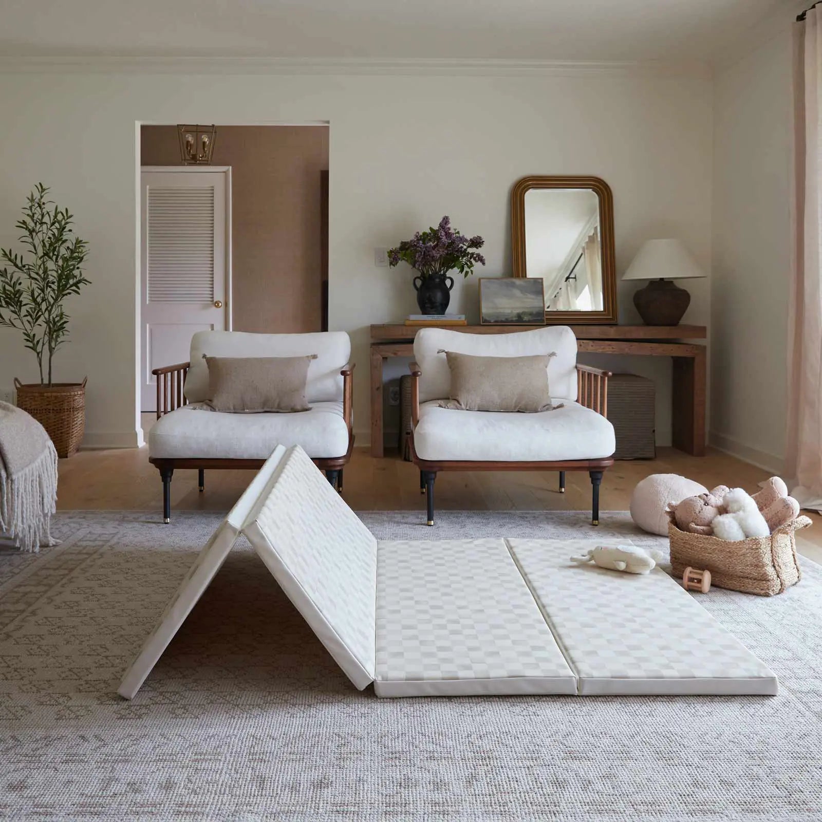 Almond neutral checker print tumbling mat shown in living room with baby girl walking across the mat toward toys and stuffed animals