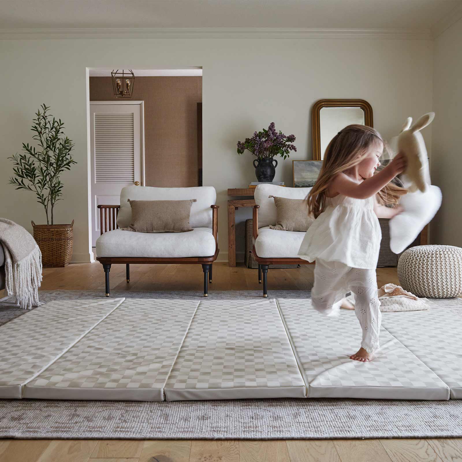 Almond neutral checker print tumbling mat shown in living room in size 5x7.5 with little girl dancing across the mat with stuffed animals in her hands