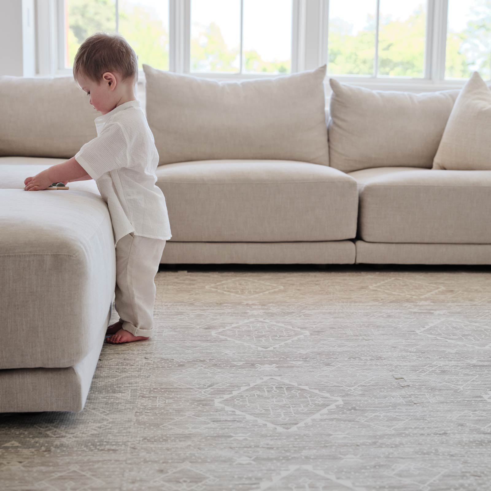 Ula driftwood neutral boho pattern play mat shown in a living room with a toddler boy playing with a toy standing against a light beige linen couch