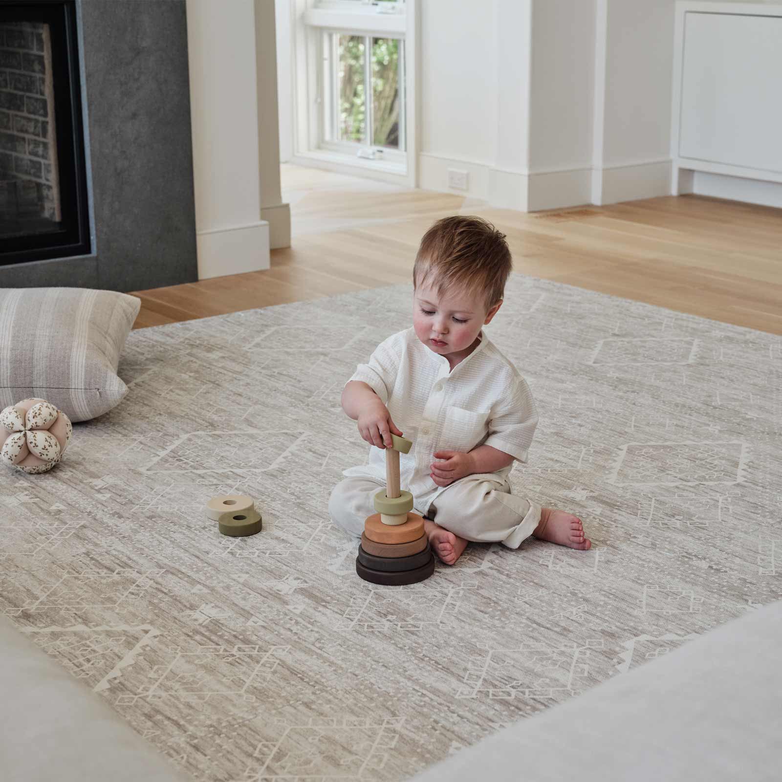 Ula driftwood neutral boho print play mat shown in a living room with toddler boy playing with a stacking toy on the mat