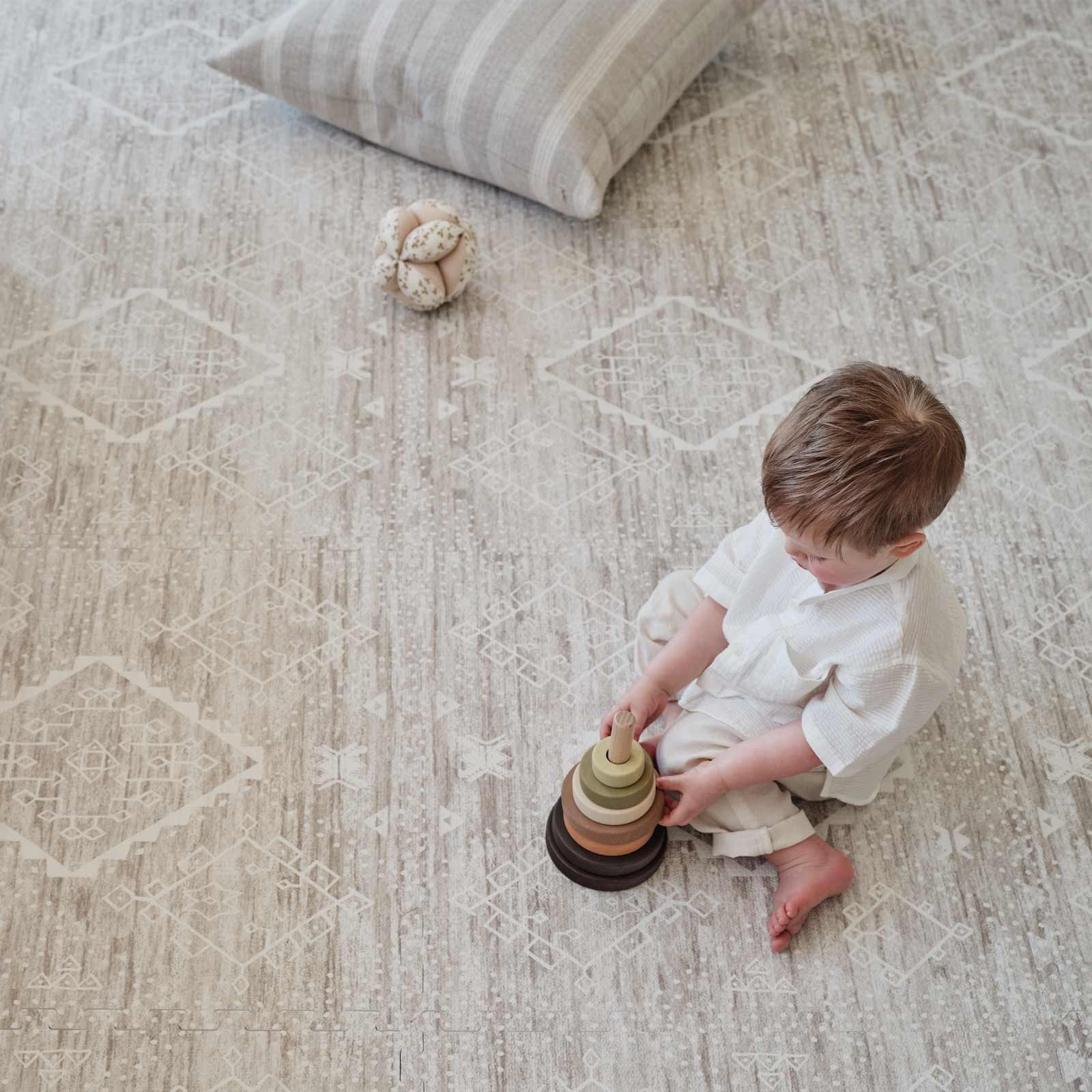 Ula driftwood beige and white boho print play mat with toddler boy sitting on the mat playing with a wooden stacking toy with a pillow and plush toy behind him