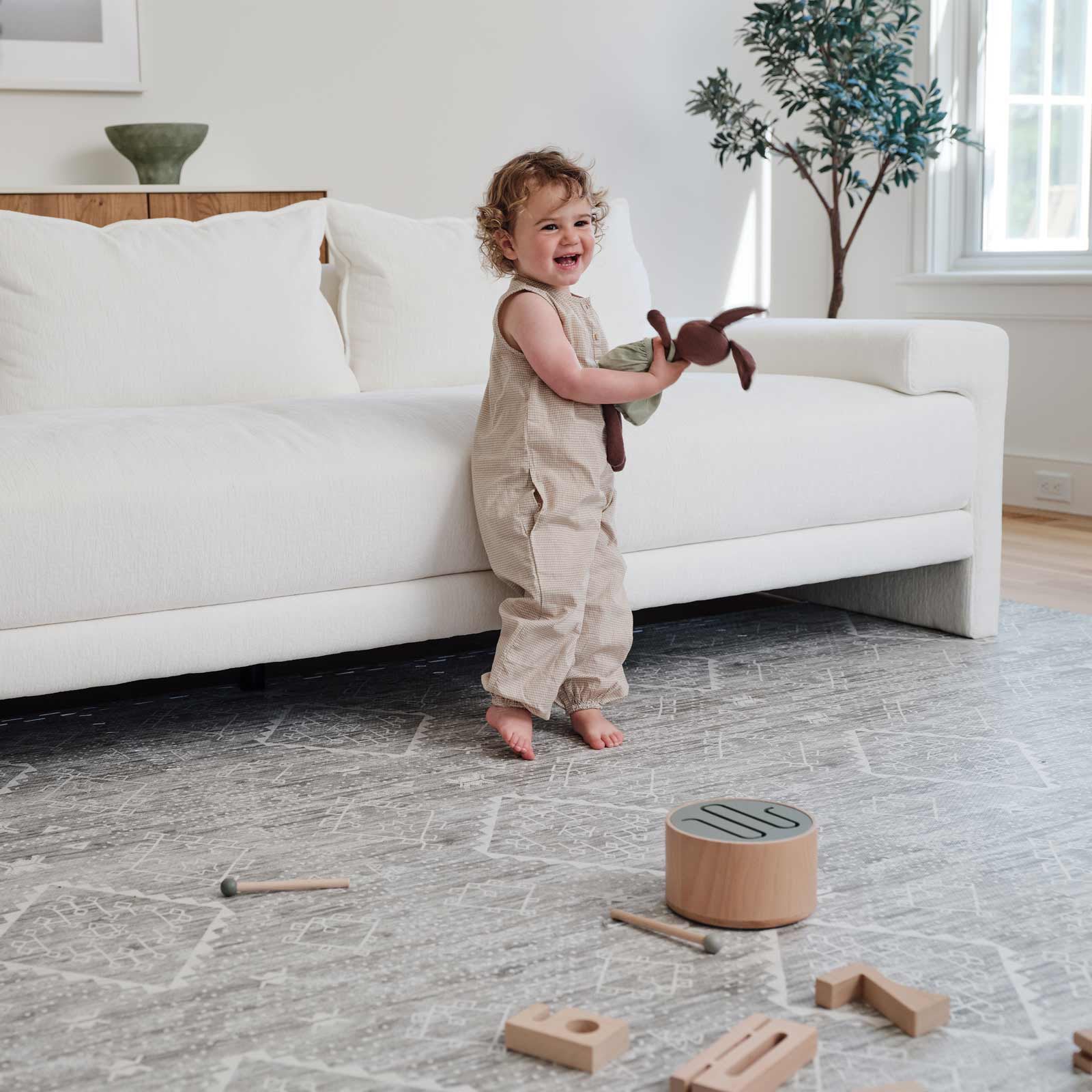 Ula willow sage green boho print play mat shown in a living room under a white couch with toddler boy holding a bunny toy with wooden blocks in front of him on the mat