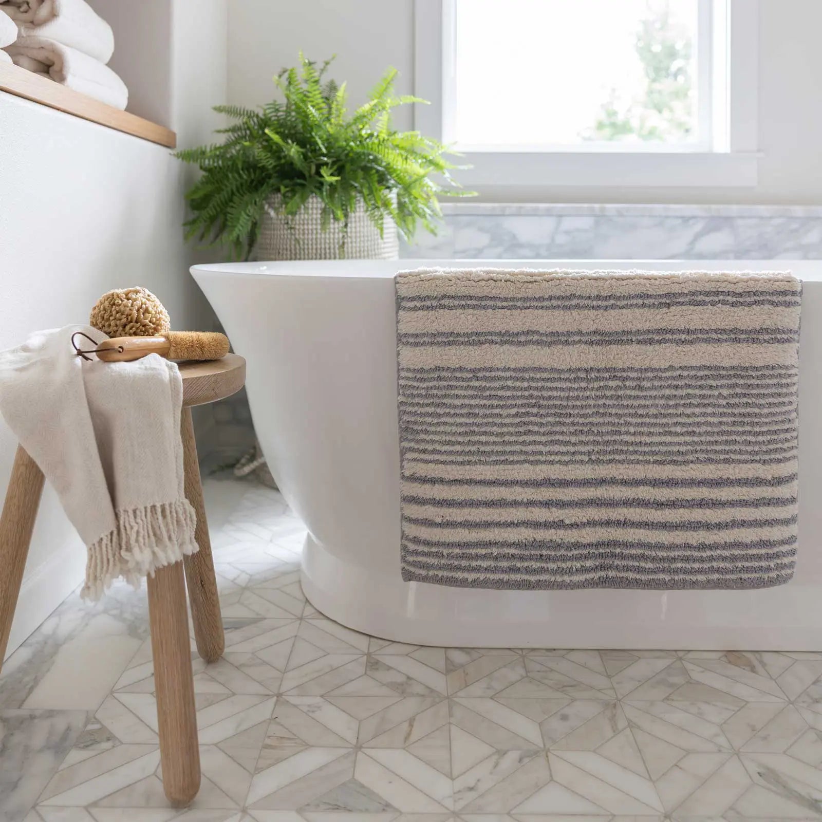 Coastal Nantucket blue and white stripe bath mat in size 21x34 hanging over the side of a bath tub with a wooden stool holding bath supplies next to the tub