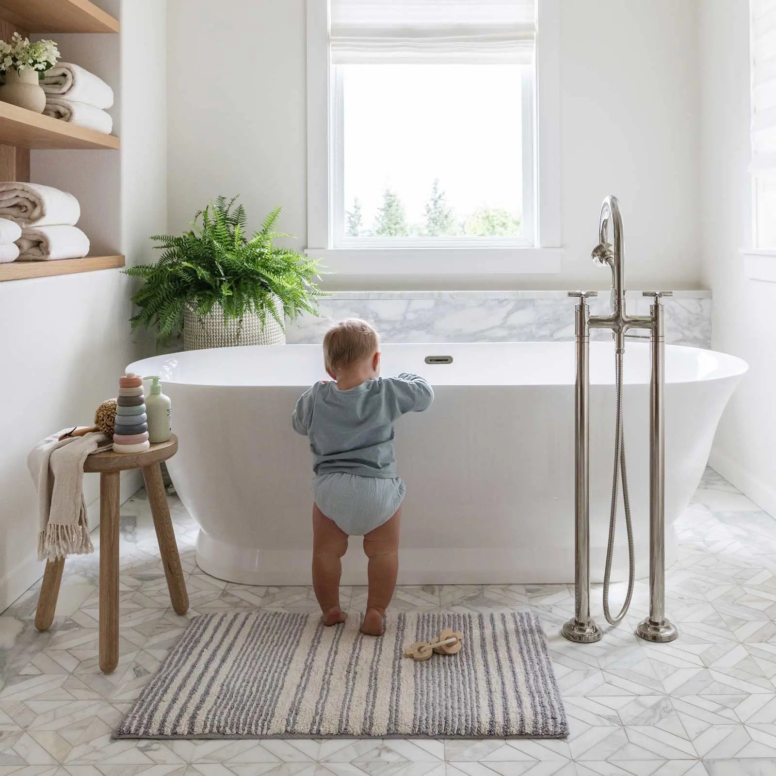 Coastal Nantucket blue and white stripe bath mat in size 21x34 shown in a bathroom in front of a tub with baby standing up on the mat pulling themselves up on the tub