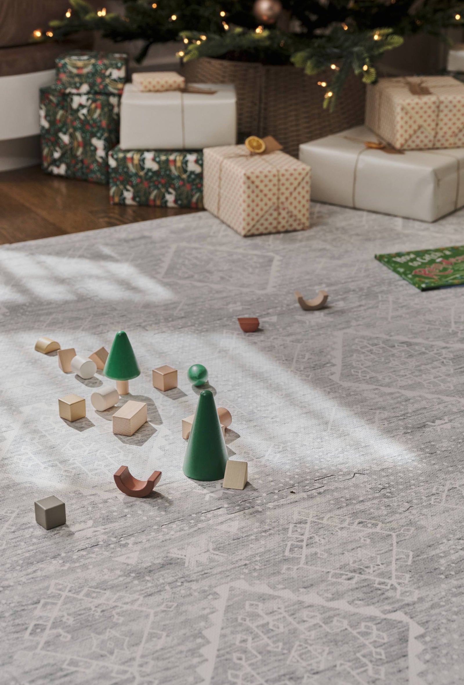 Ula gray boho print play mat with wooden blocks laid out on the mat. Lit up christmas tree with wrapped gifts underneath in the background